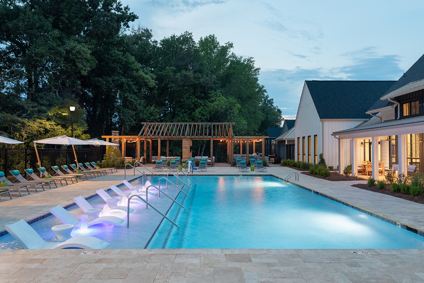 Outdoor swimming pool with white in-water lounge chairs beside a modern farmhouse-style clubhouse.